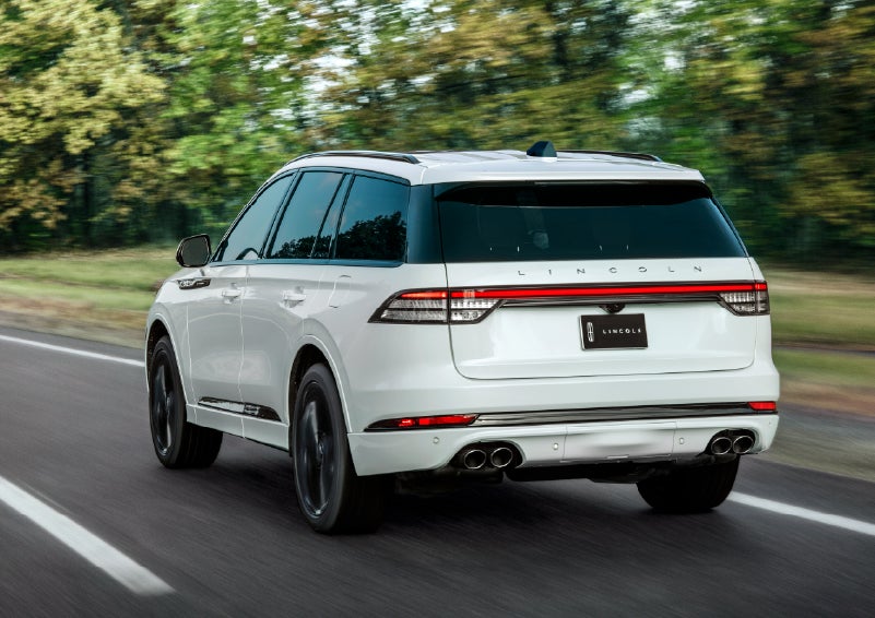 A white 2026 Lincoln Aviator® SUV is being driven away down a road lined with colorful trees. | Lincoln North Miami in North Miami FL