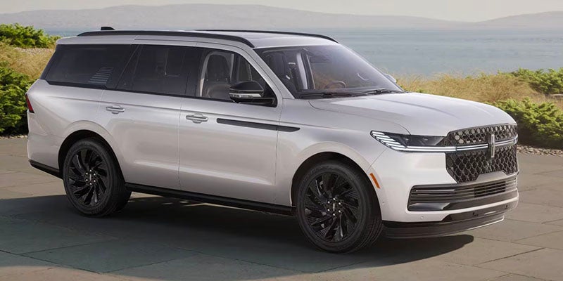 Silver Lincoln Navigator SUV with black wheels parked outdoors, with water and greenery in the background.