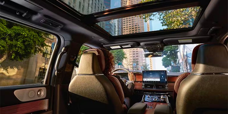 Inside a car with a panoramic sunroof, showing the driver, large infotainment screen, and city buildings through the windows.