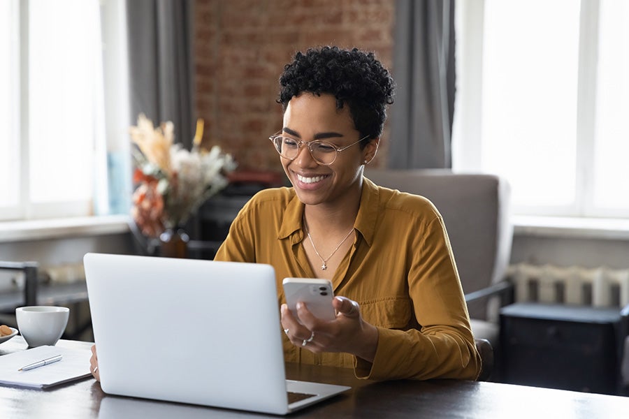 Smiling Black woman with glasses, short curly hair, using a laptop and phone at a desk.
