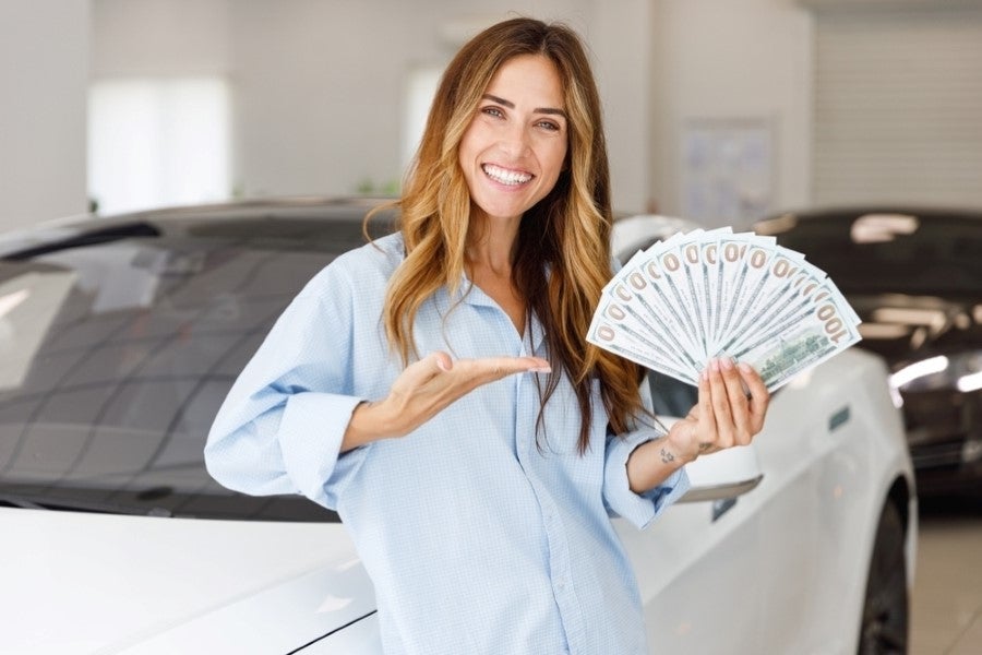 Person standing beside a vehicle in a dealership showroom, holding a fan of cash and gesturing toward the car